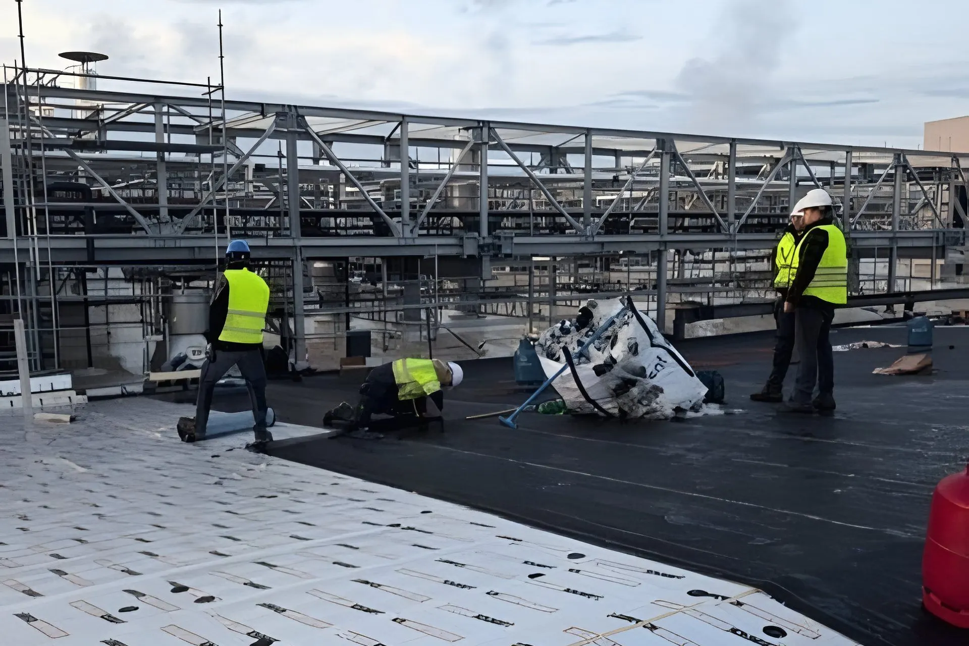 Toiture plate industrielle en chantier : à l'avant des panneaux d'isolation PIR imprimés, derrière l'étanchéité bitumineuse noire et quelques personnes en gilet fluo près d'une structure technique en acier