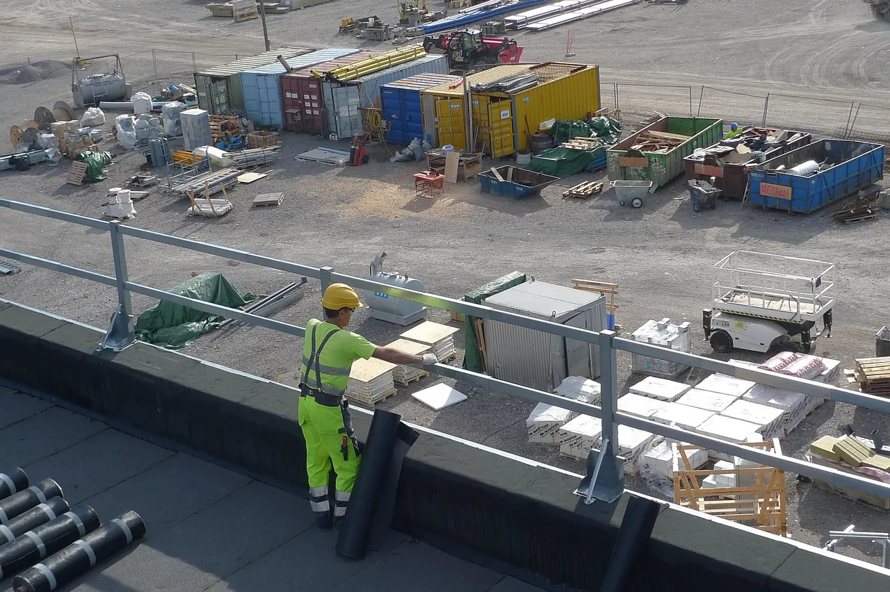 Un ouvrier en tenue fluo et casque au bord d'une toiture industrielle en chantier, à côté de rouleaux de bitume, avec en contrebas les conteneurs et le matériel du chantier